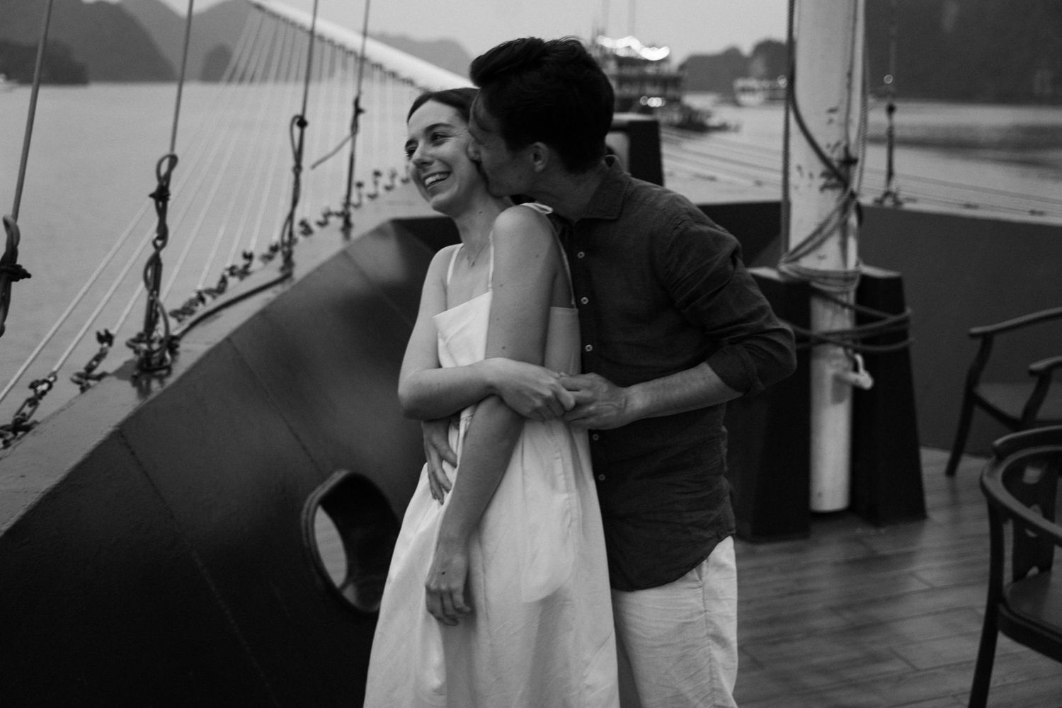 Black and white photo of couple embracing on boat deck with nautical elements visible.