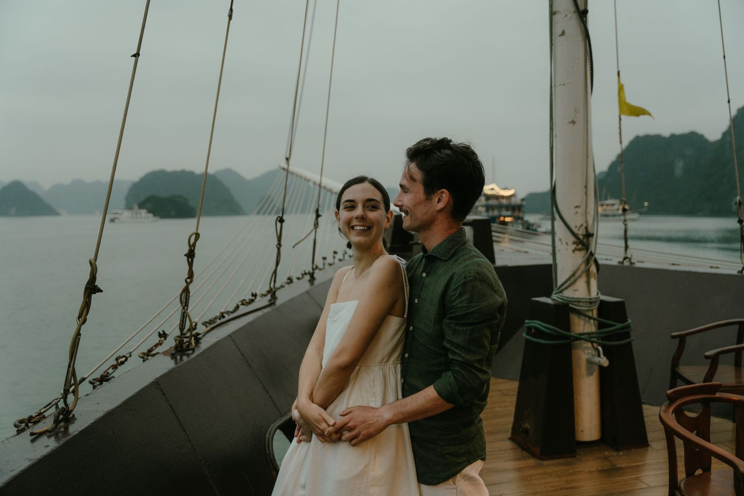 Couple sharing tender moment on sailing vessel with dramatic mountain landscape behind them.