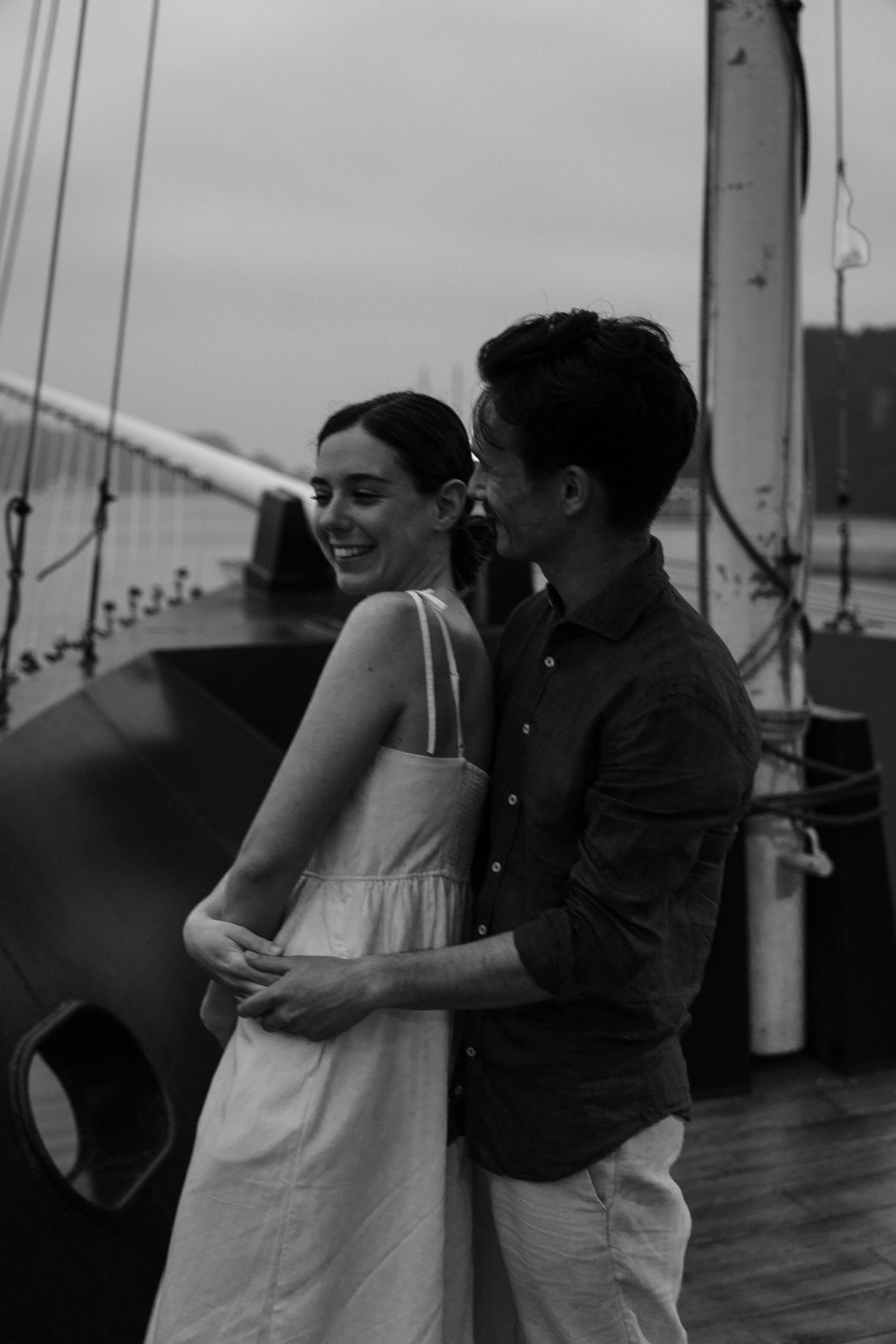 Black and white candid moment of couple laughing together on boat deck with mast visible.
