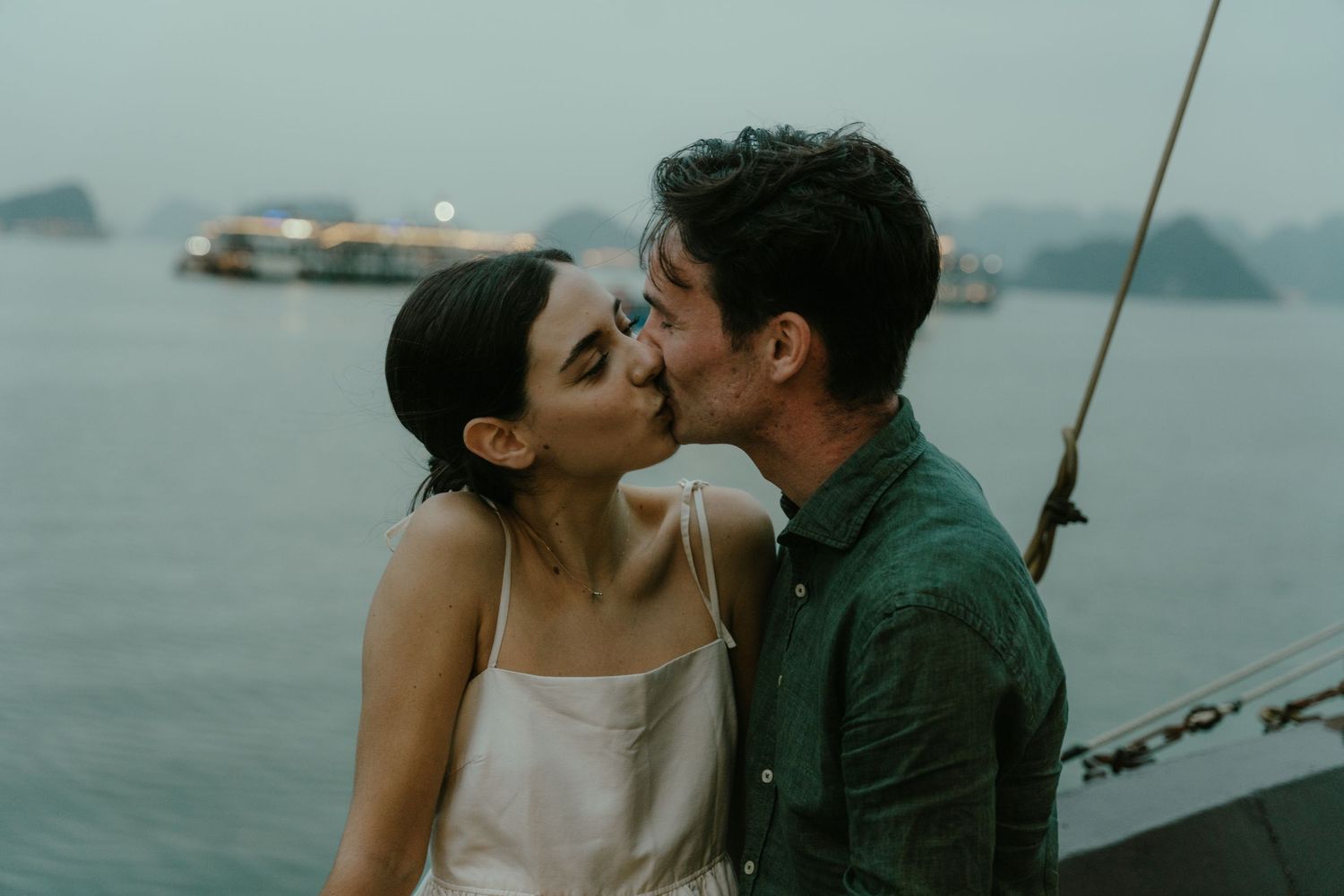 Intimate kiss shared between couple on boat with misty mountains and water in background.