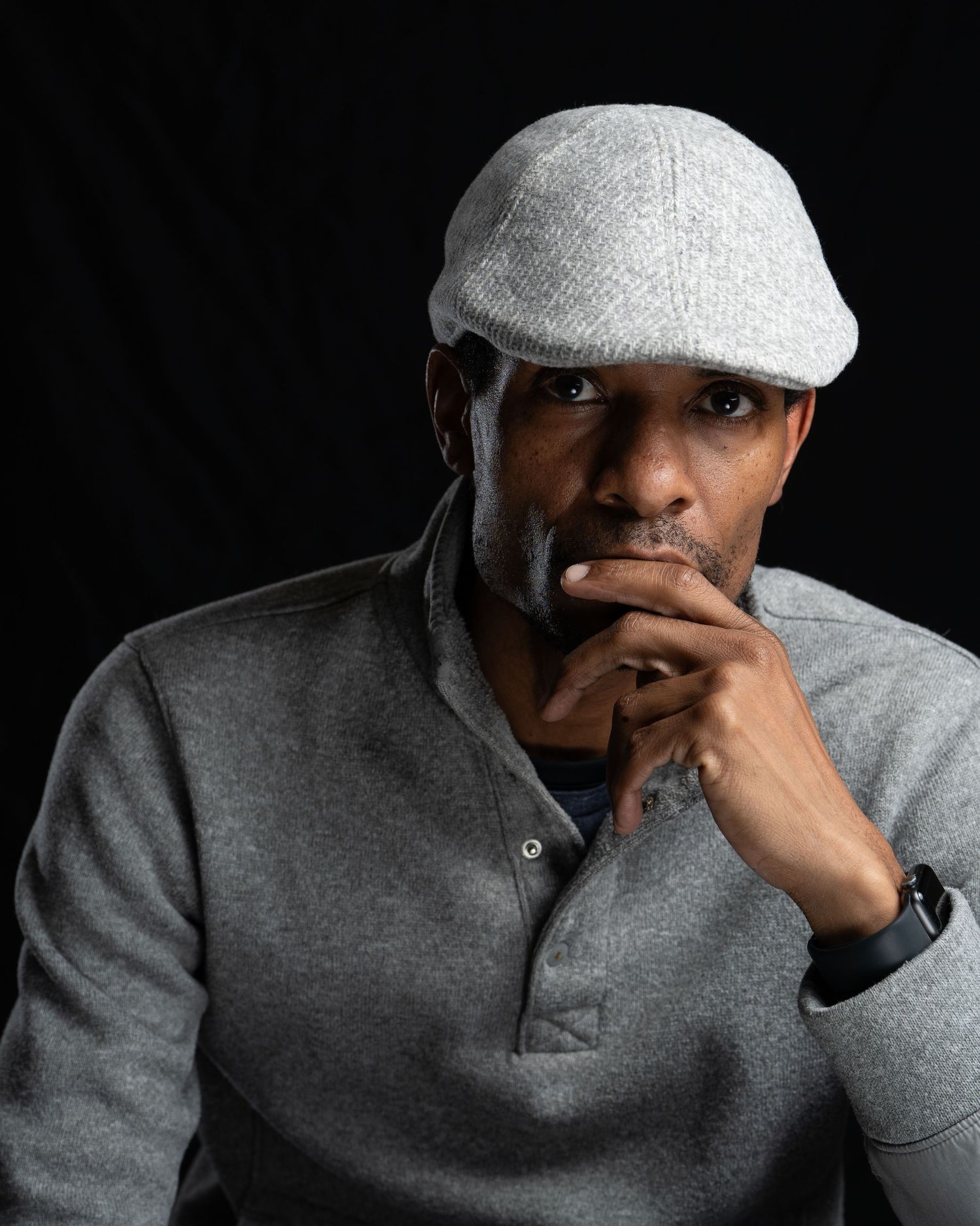 Thoughtful pose in gray clothing and cap against dark backdrop.