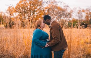 A couple stands together in a golden autumn field during sunset while wearing brown and teal colored outfits.