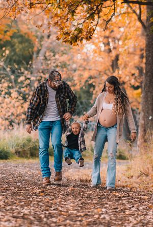 Family walks together on fallen autumn leaves along a wooded path during golden hour with warm orange foliage above.