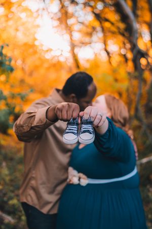 A pair of tiny baby shoes held up against an autumn forest backdrop with warm orange foliage in the background.