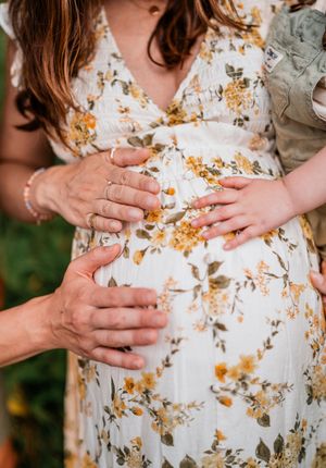Multiple hands rest lovingly on a pregnant belly covered in a white and yellow floral dress in an outdoor setting.