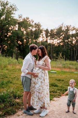 A couple shares tender moments in a grassy field at sunset while their toddler plays in the background.