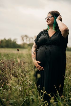 A person in a black dress stands in a wildflower meadow during a maternity photoshoot on an overcast day.