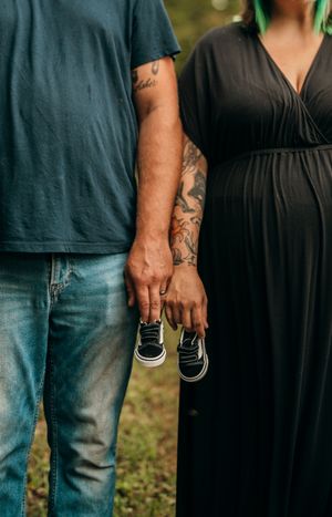 A couple holding hands outdoors, showing tattoos on their arms and wearing casual clothing in a natural setting.