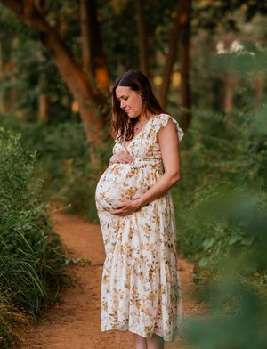 A pregnant woman in a floral dress walks along a wooded trail during golden hour sunlight.