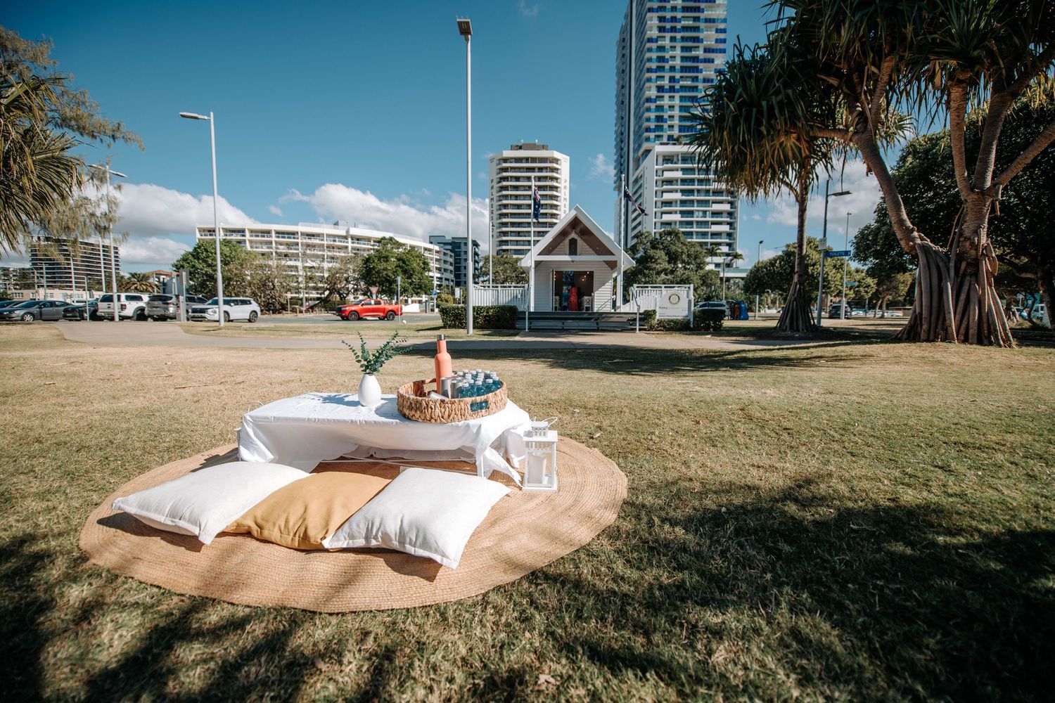 Picnic setup with pillows and drinks on grass with white chapel and city buildings in background.