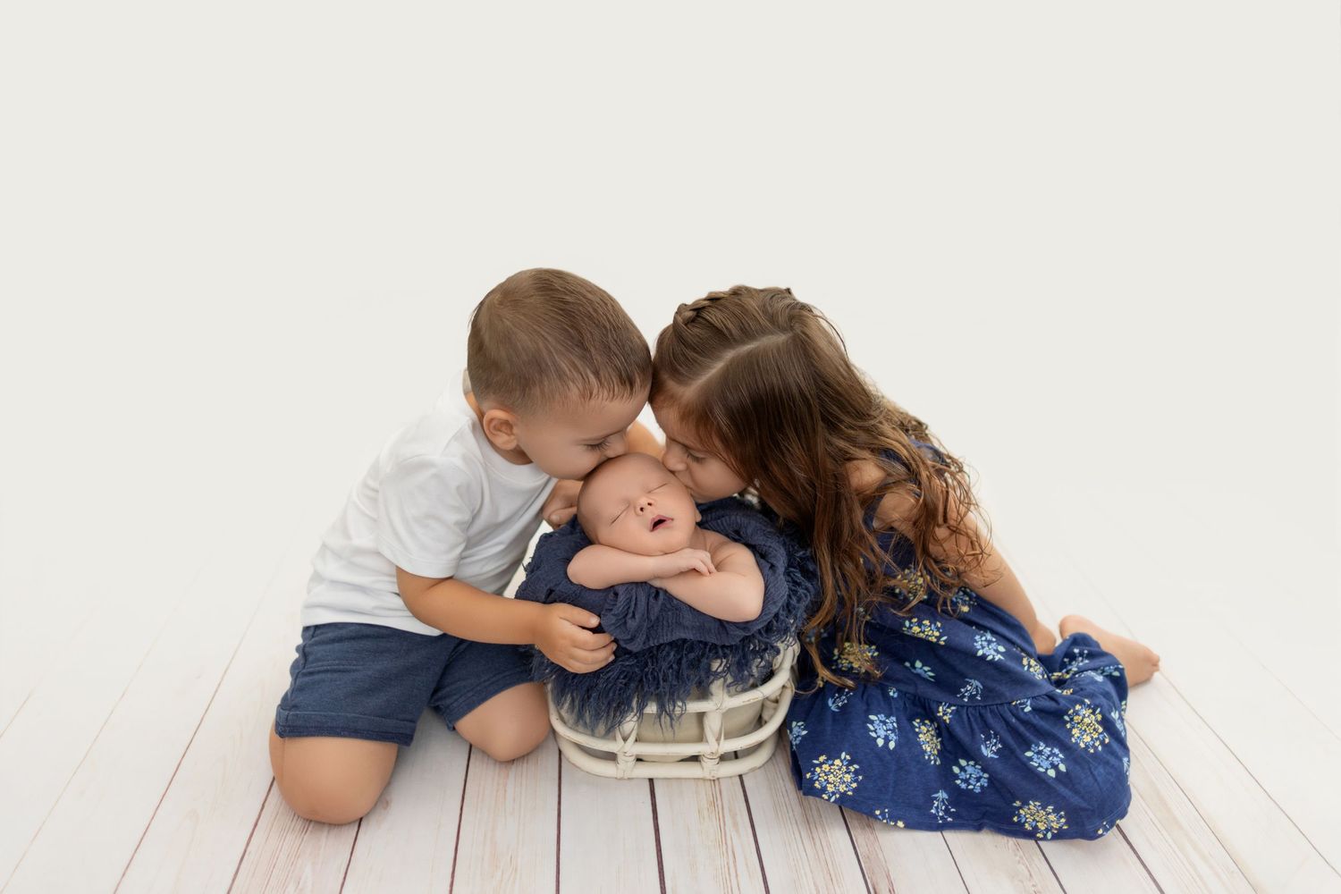 Two small children leaning in to kiss a baby lying on a white wooden floor.