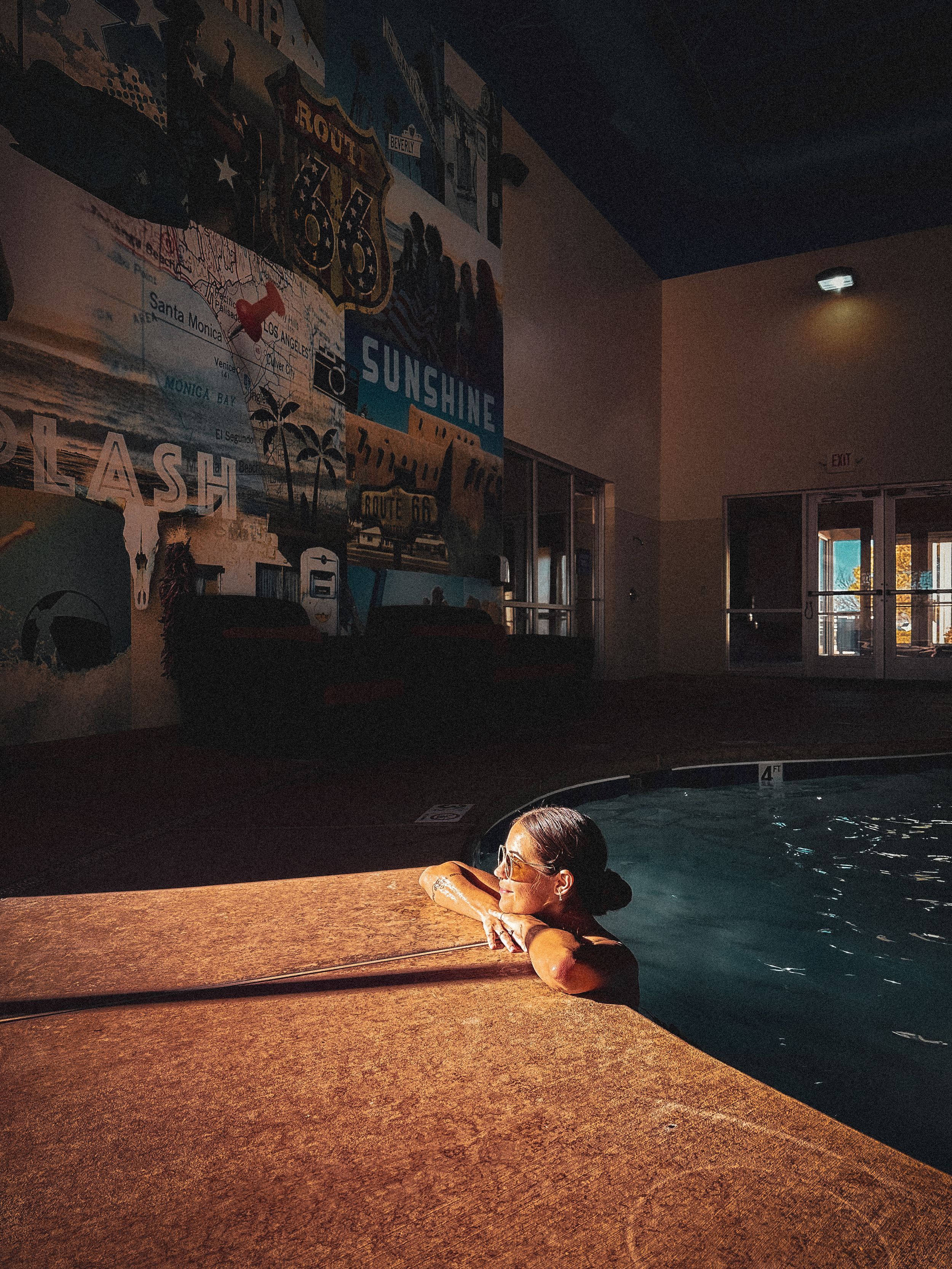 Person relaxing in an indoor pool beside a colorful Route 66 sunshine mural at night.
