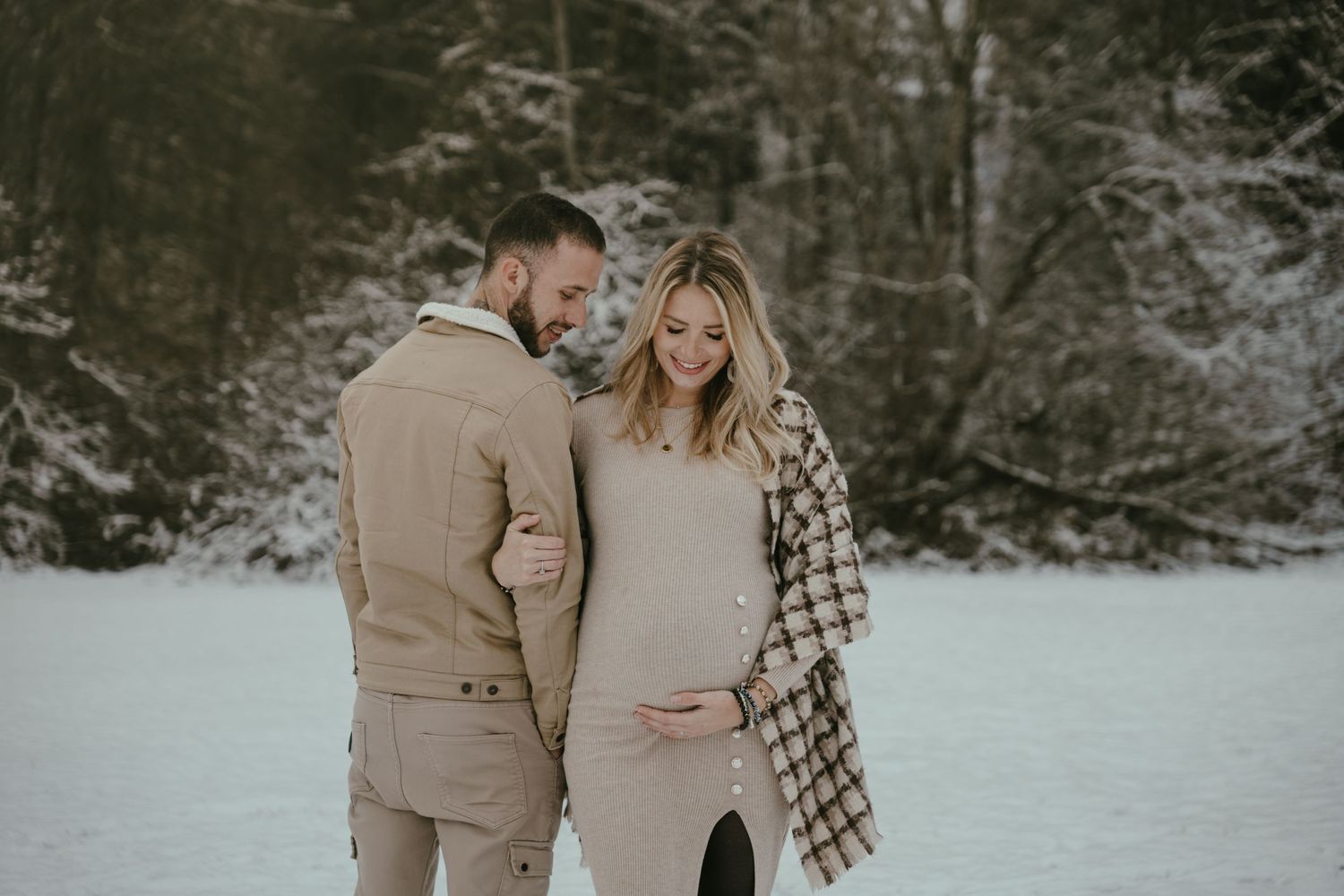 A couple stands together in a snowy winter scene wearing coordinating beige and plaid outfits.