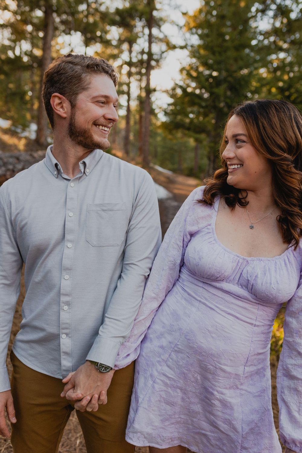 A couple shares a joyful moment while holding hands in casual attire against a forest backdrop.