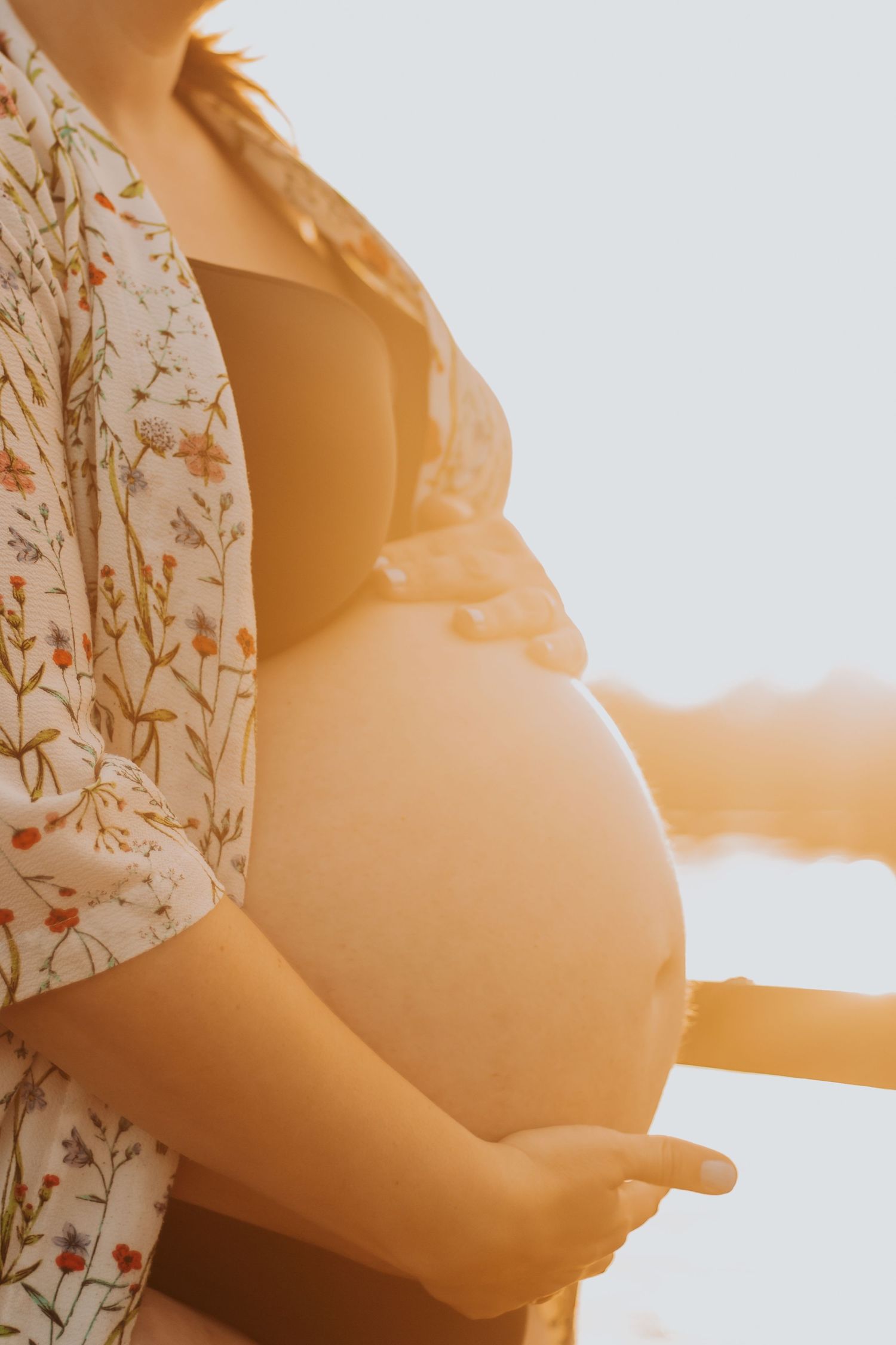 Close-up silhouette of a pregnant belly in floral dress against golden sunlight.