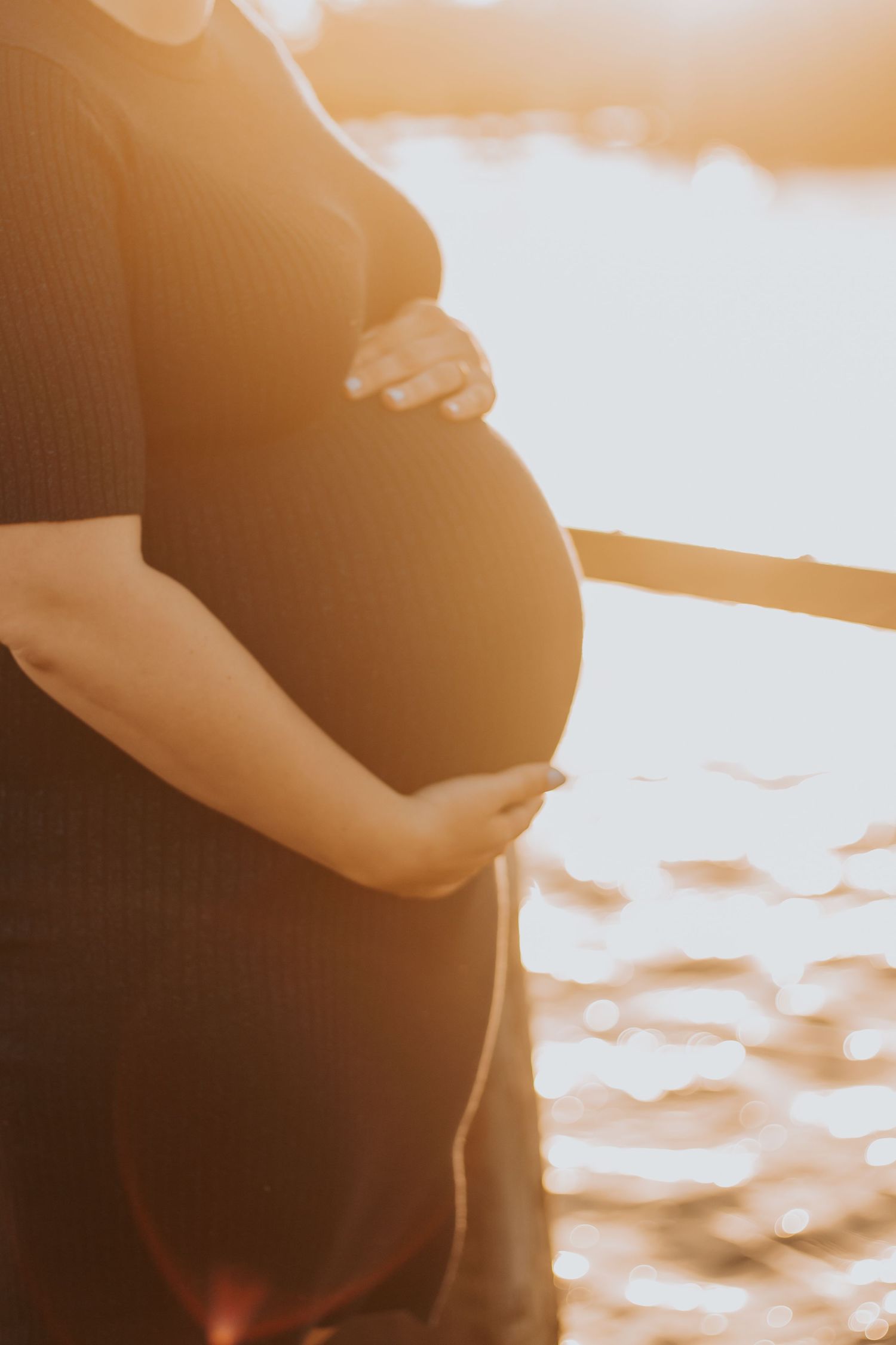 Silhouetted baby bump being cradled by hands against a sunset ocean backdrop.
