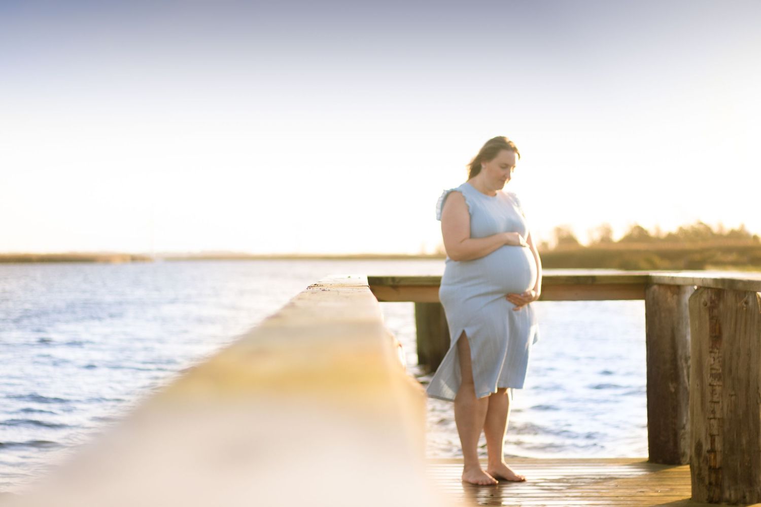 A pregnant silhouette stands peacefully on a pier overlooking the ocean at sunset.