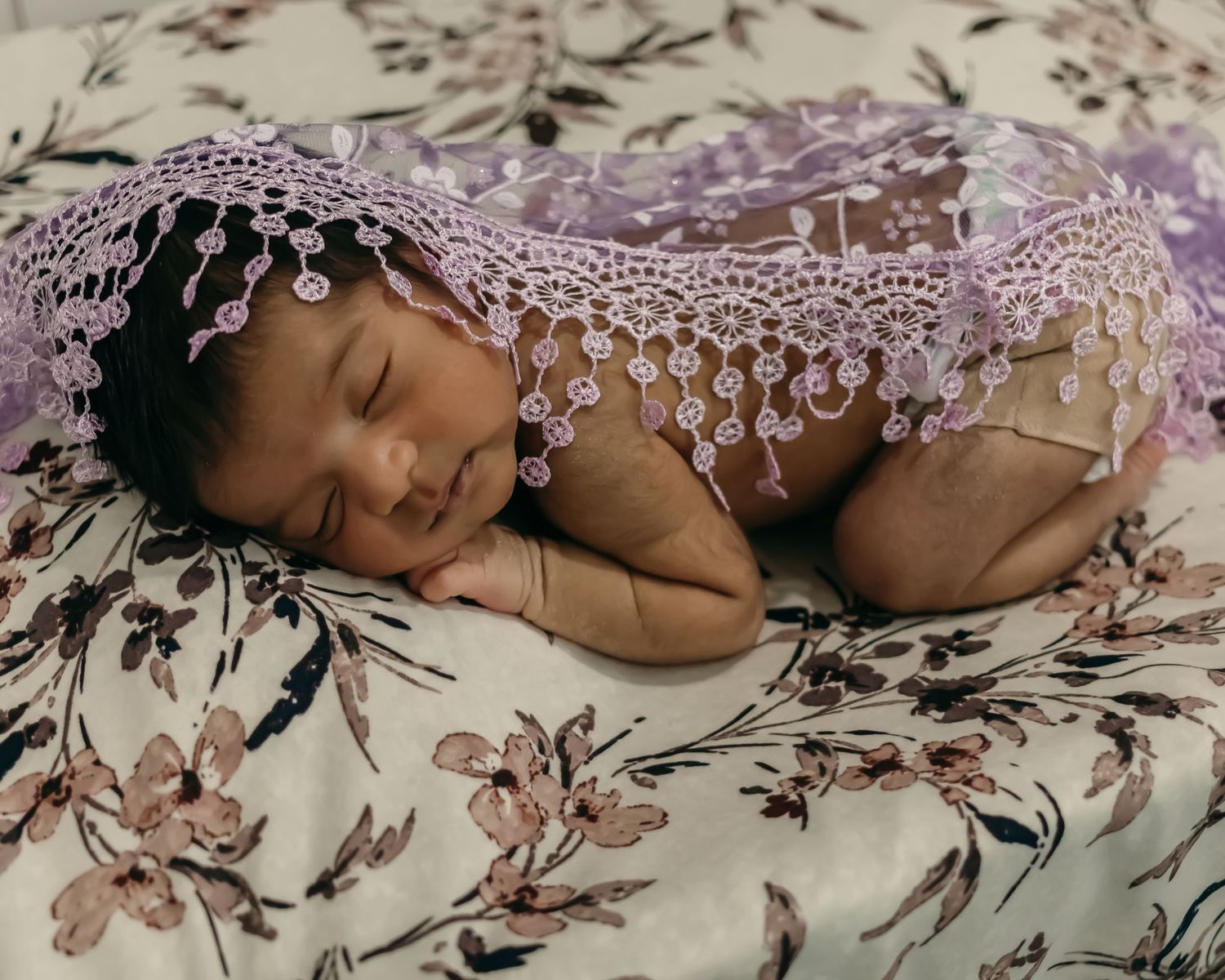Sleeping infant wrapped in delicate purple lace on a floral pattern fabric.
