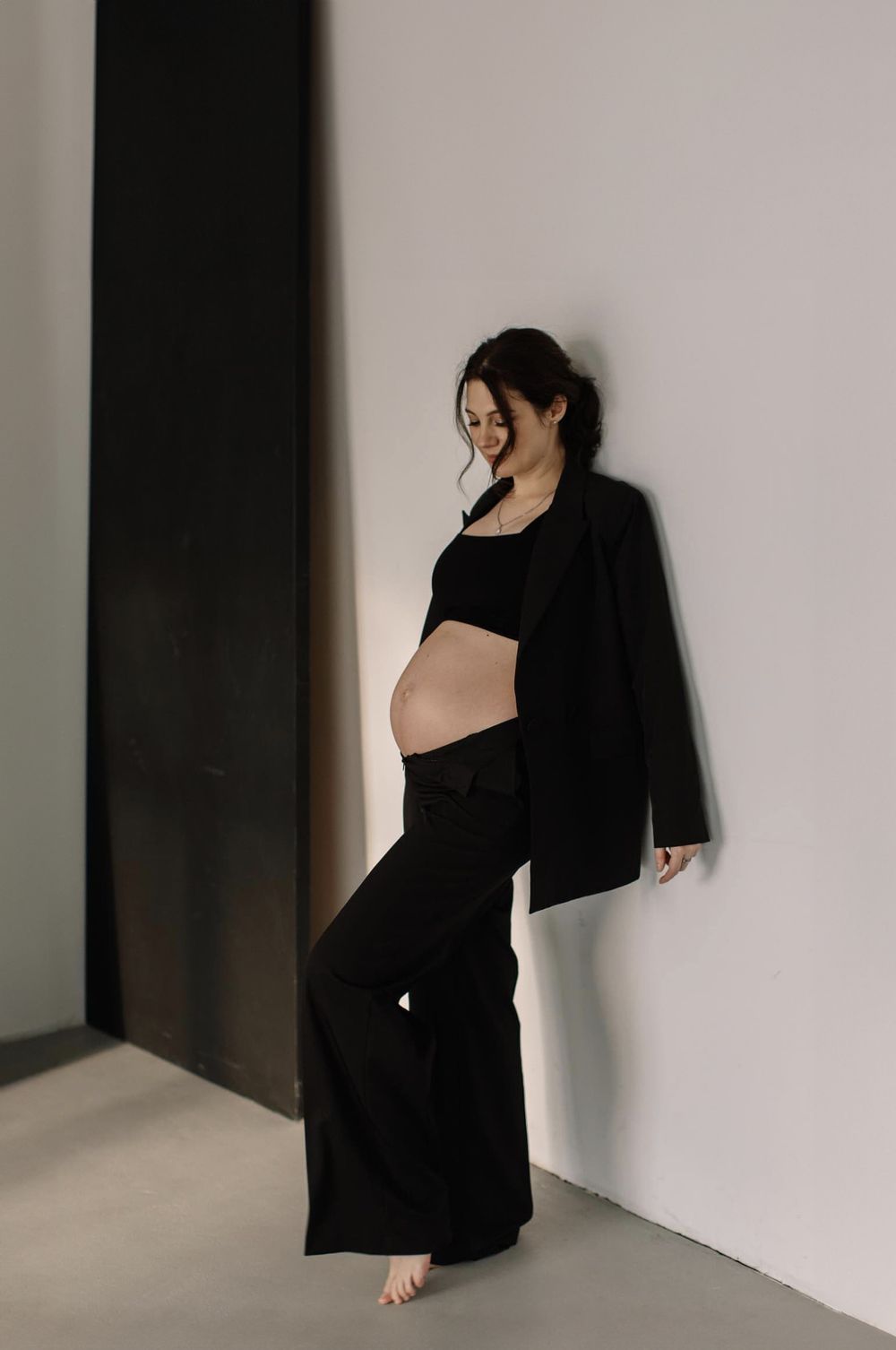Minimalist maternity photo featuring all-black ensemble against white wall.