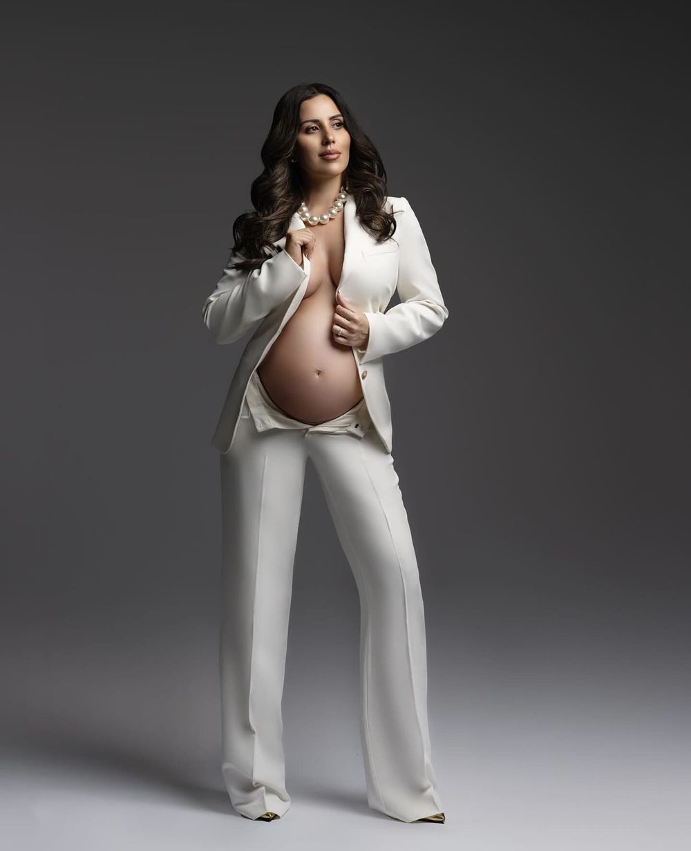 Modern maternity portrait in white pantsuit against dark studio background.
