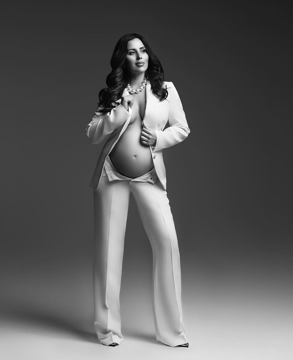 Black and white maternity portrait in elegant white pantsuit against studio backdrop.