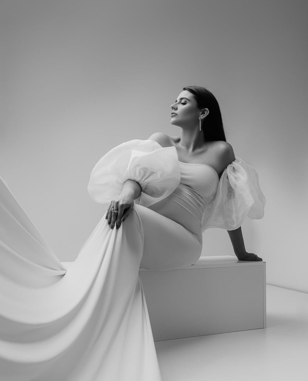 Black and white studio portrait with dramatic white gown and puffy sleeves while sitting on cube.