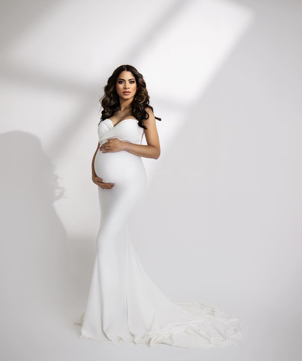 Elegant maternity portrait in fitted white gown with dramatic train against white studio backdrop.