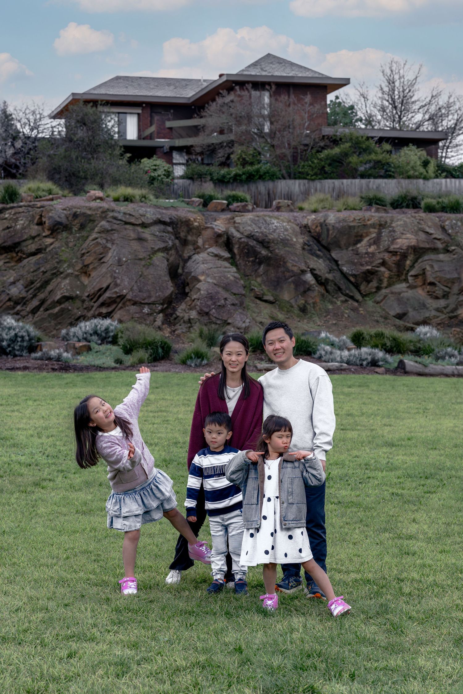 Asian family of five posing on green lawn with rocky hillside and modern home in background.