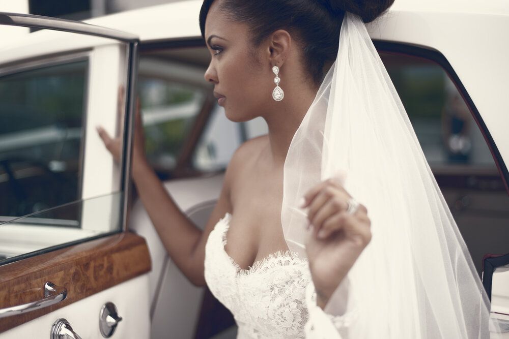 Elegant bride in lace gown and veil standing beside a vintage white car.