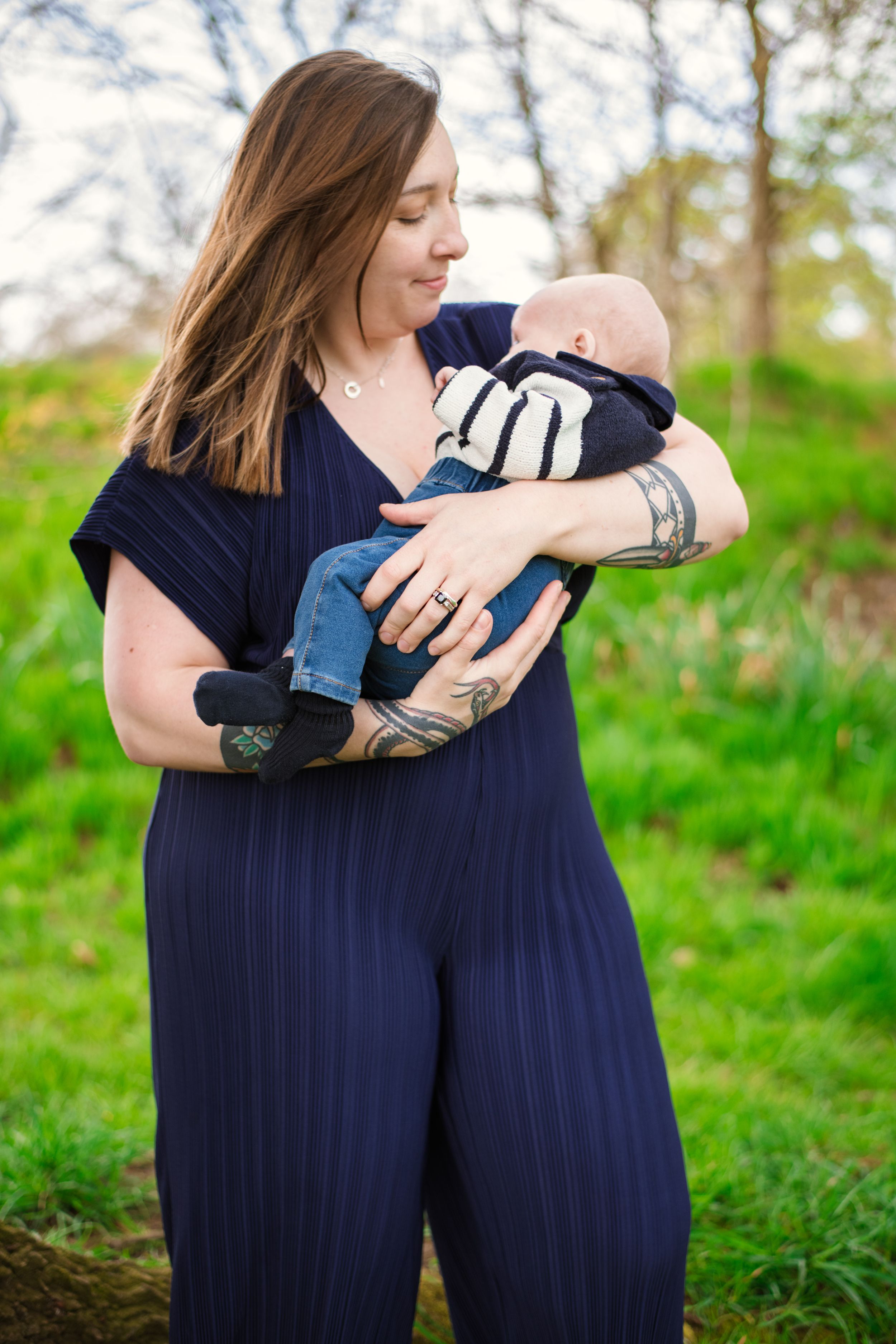 Woman in navy jumpsuit holding infant outdoors in natural setting.