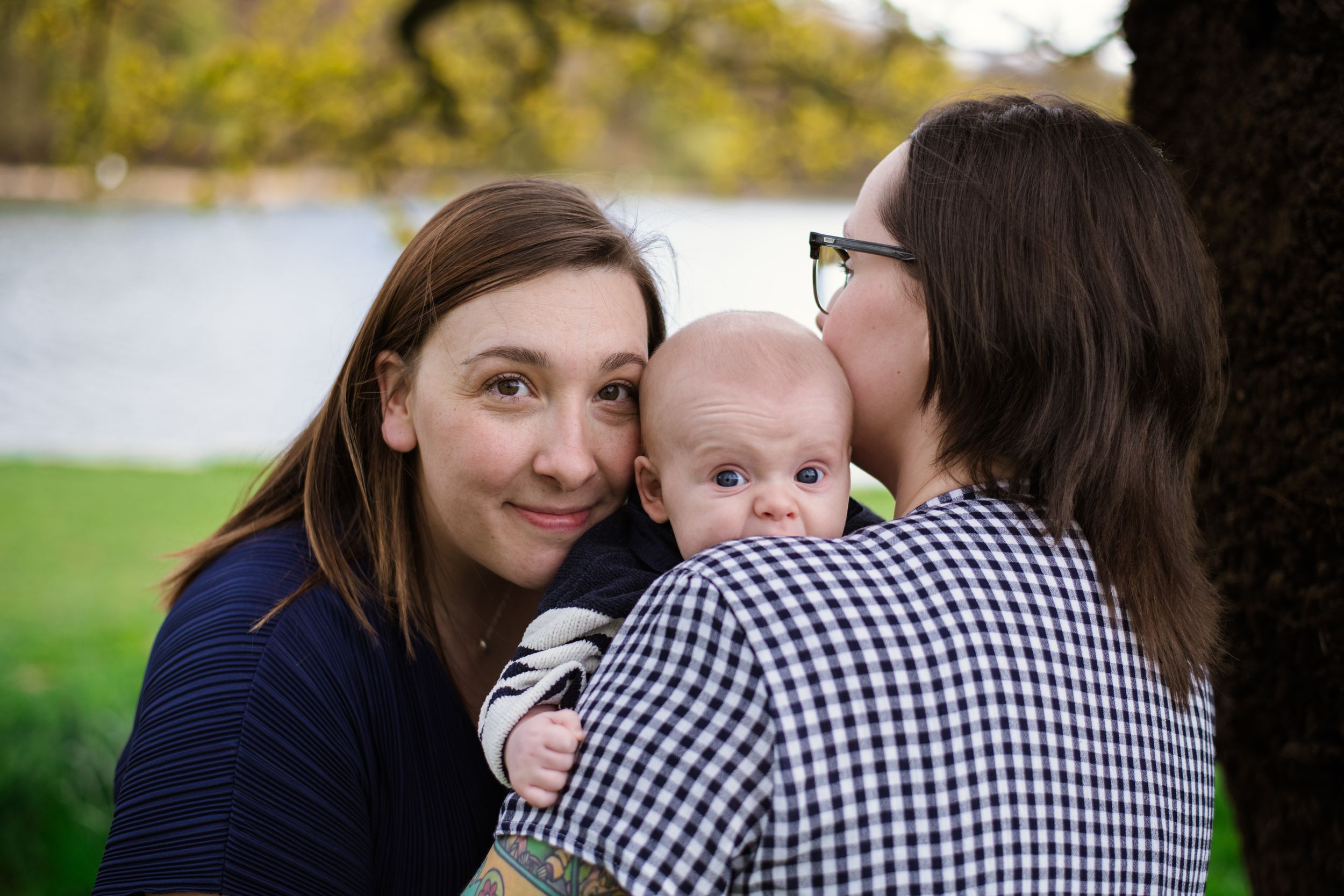 An intimate family moment outdoors by a lake with a baby being held between two people.