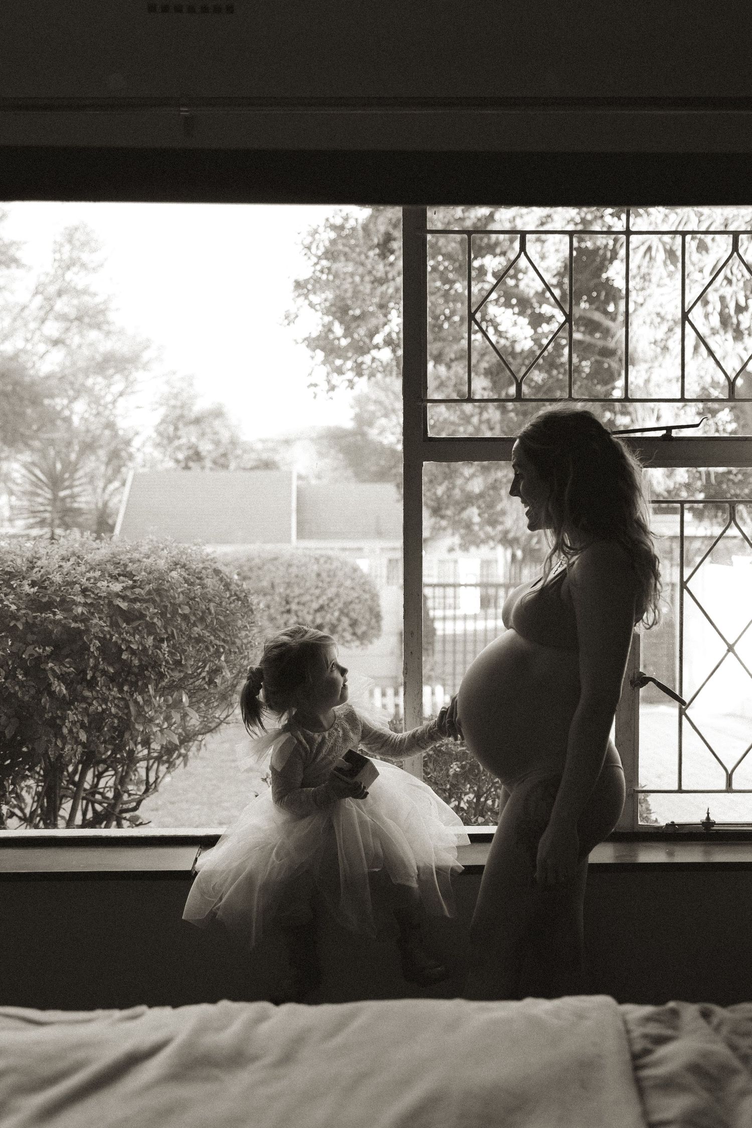 A black and white portrait of a pregnant woman in a white top leaning back against a wall.