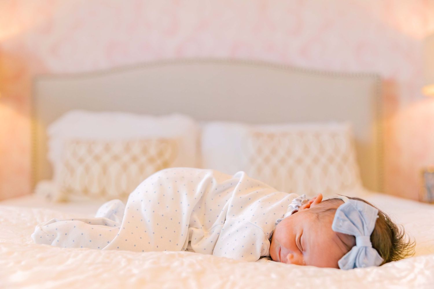 A sleeping newborn in white with a bow headband rests peacefully on cream bedding.