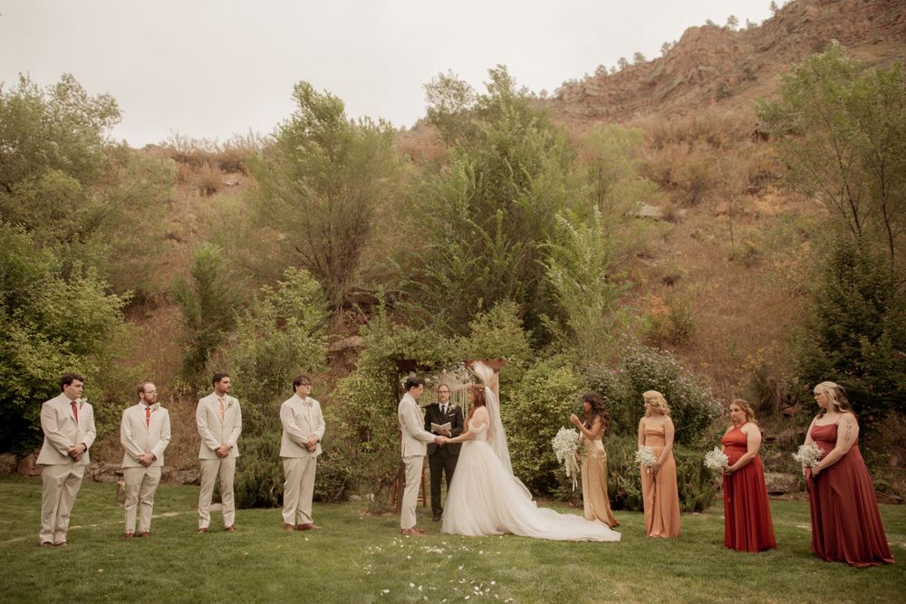 Wedding ceremony takes place in a garden setting with mountain backdrop and bridal party in burgundy and tan.