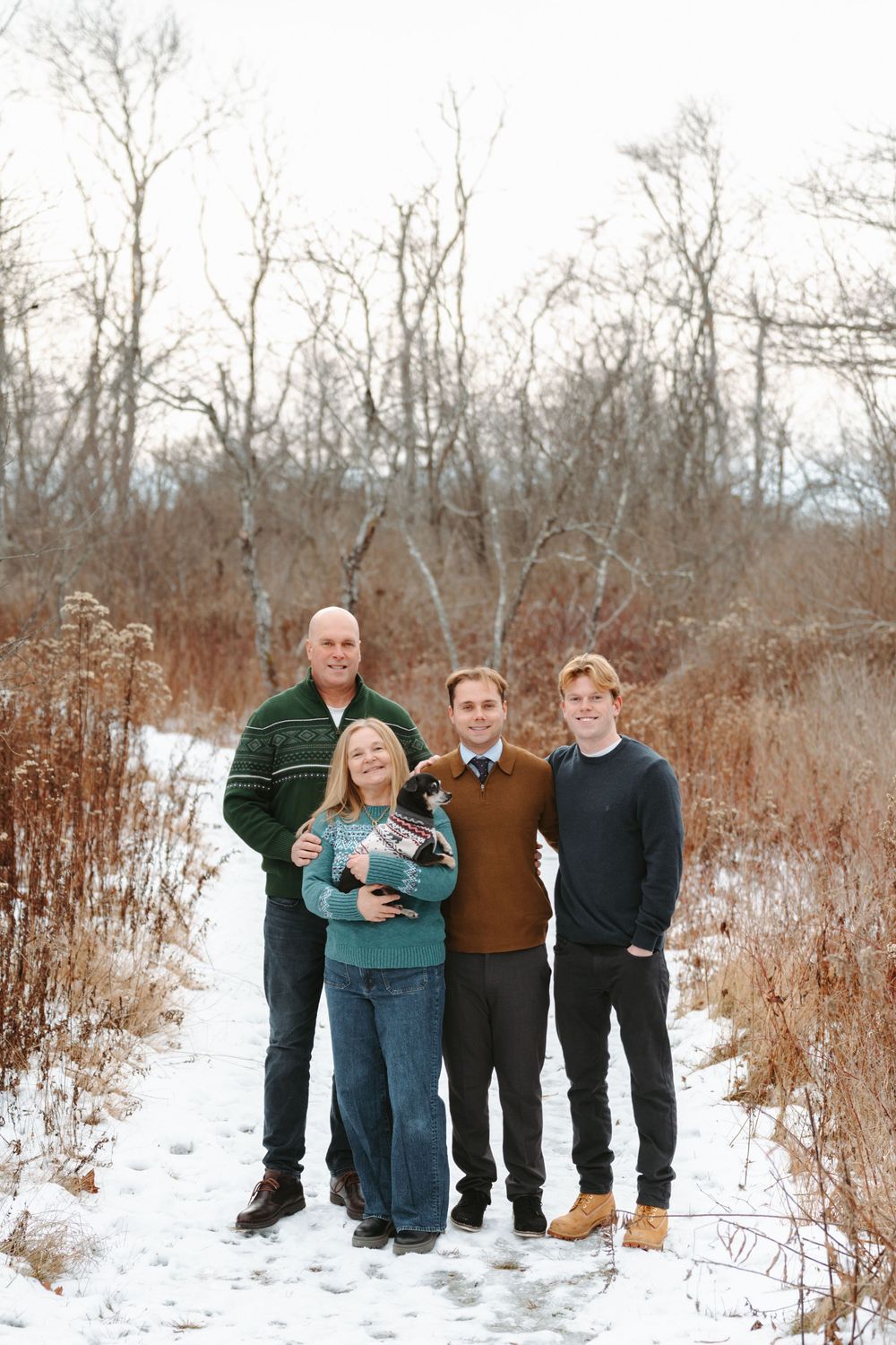 A group of people stand together on a snowy winter trail surrounded by bare trees and brush.