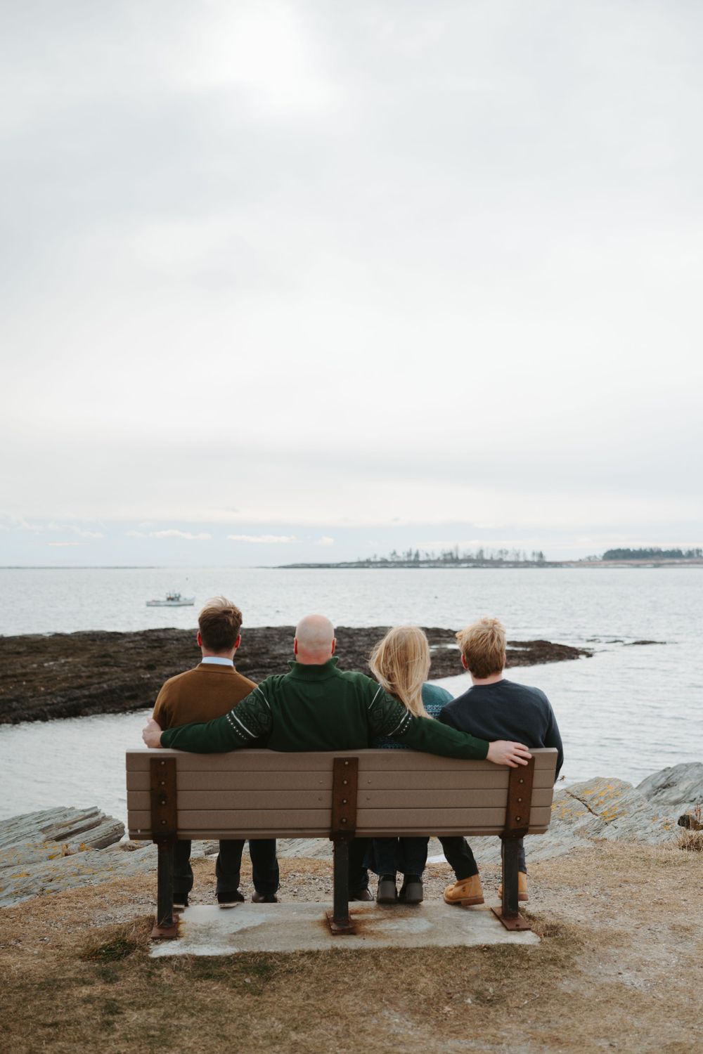 Four people sit together on a wooden bench overlooking a misty coastline and distant city skyline.