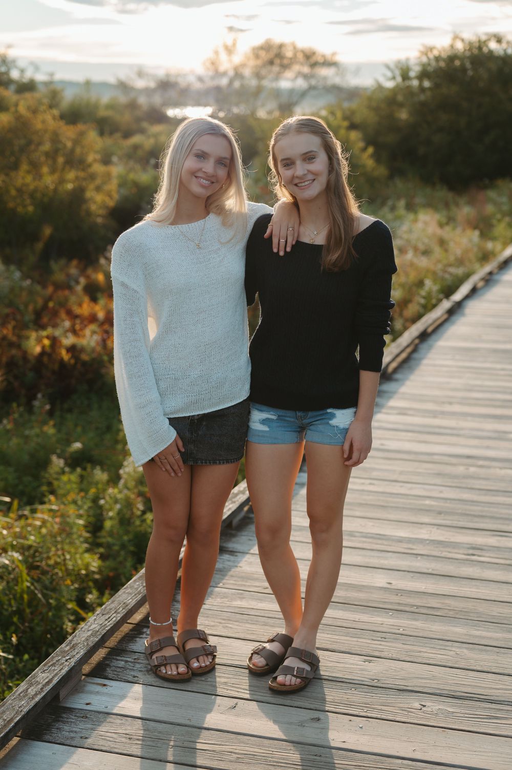 Two people in shorts and sandals stand together on a wooden boardwalk during golden hour sunset.