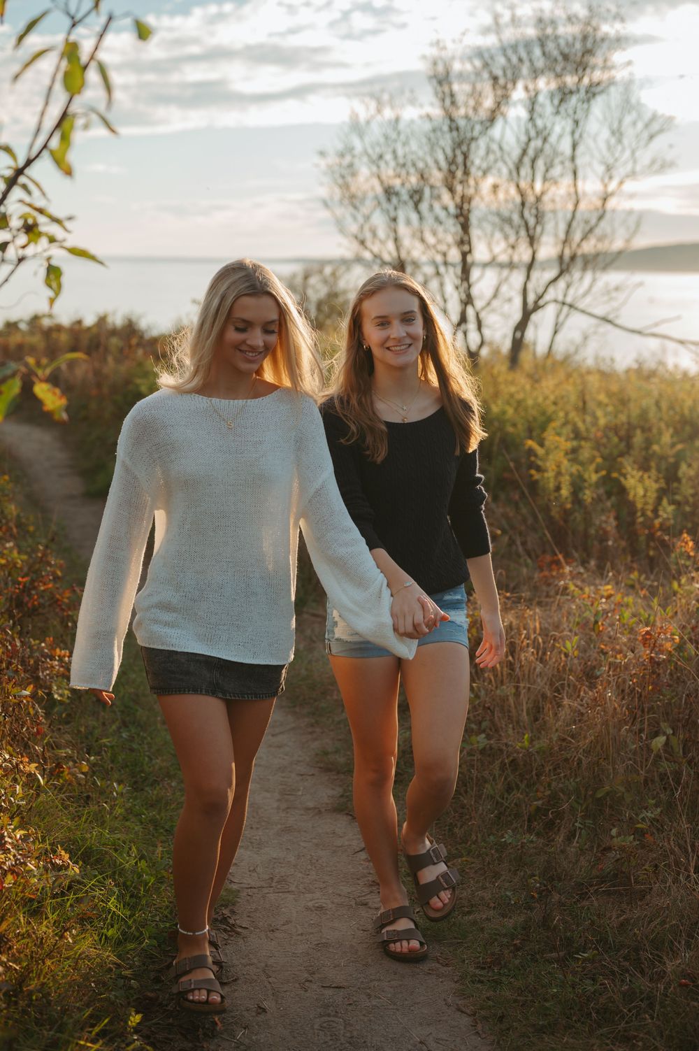 Two friends walking together on a nature trail at sunset wearing casual summer outfits.