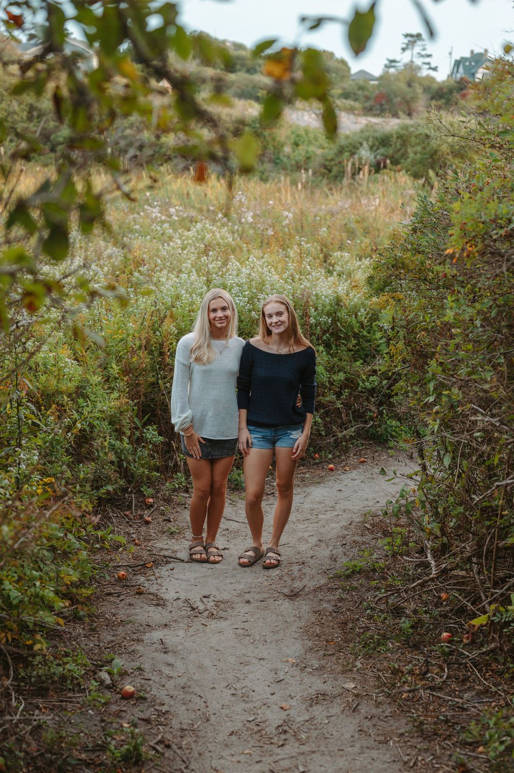 Two friends standing together on a dirt hiking trail surrounded by lush greenery and foliage on a sunny day.