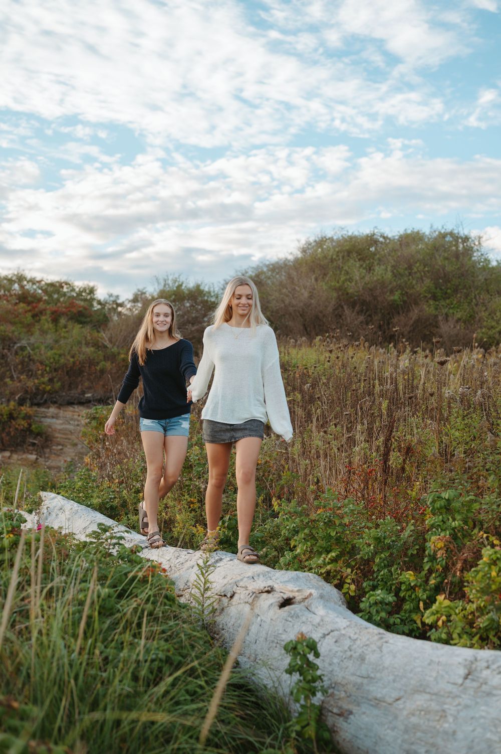 Two people exploring a coastal path while walking on a fallen tree trunk on a sunny day.