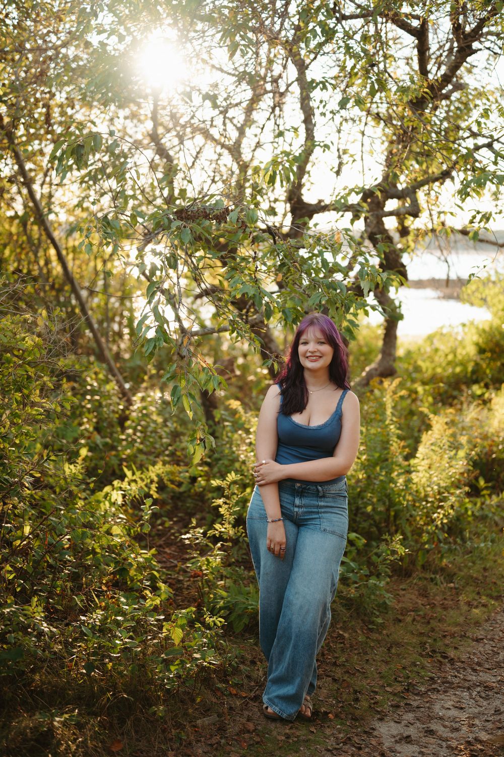 A person poses casually in blue jeans against a sunlit natural backdrop with trees and foliage during golden hour.