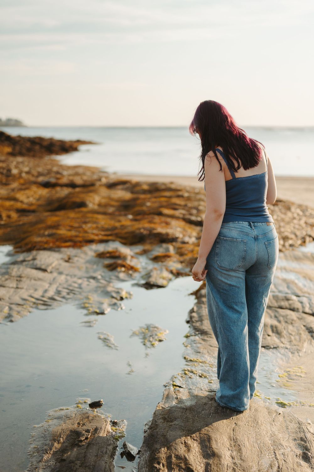 Person in denim overalls stands on rocky coastal shore during golden hour sunset by the ocean.