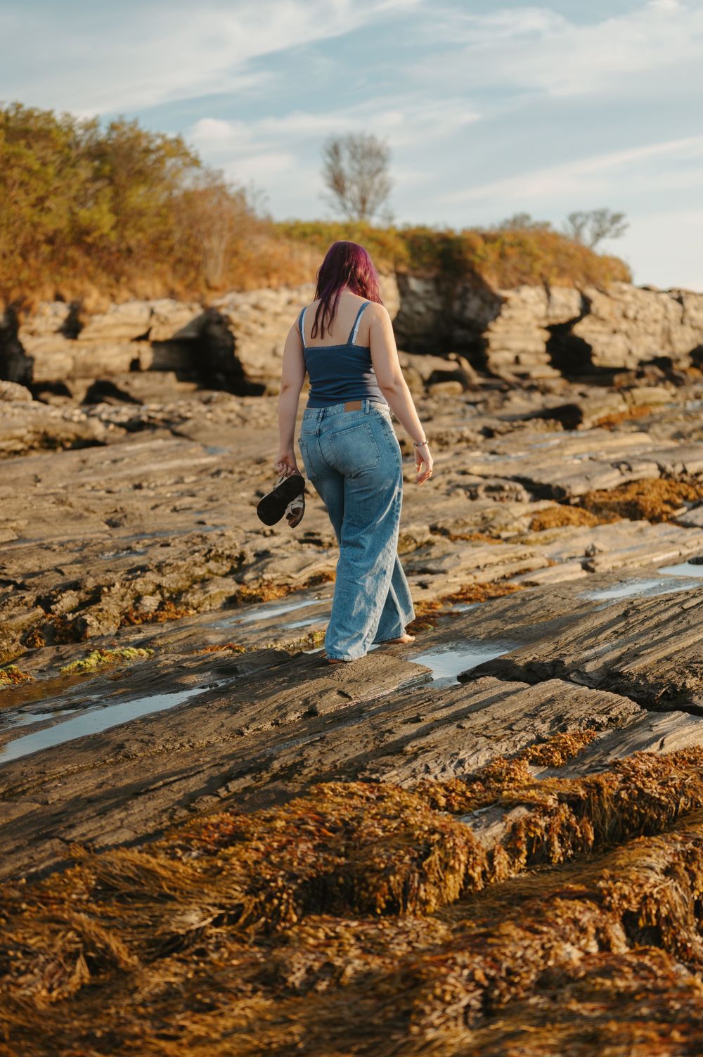 Person in blue jeans exploring rocky coastal terrain during golden hour with autumn foliage in background.