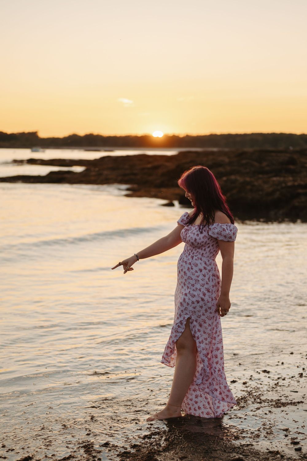 A figure in a floral dress walks along a rocky beach shoreline at sunset with golden light reflecting on the water.