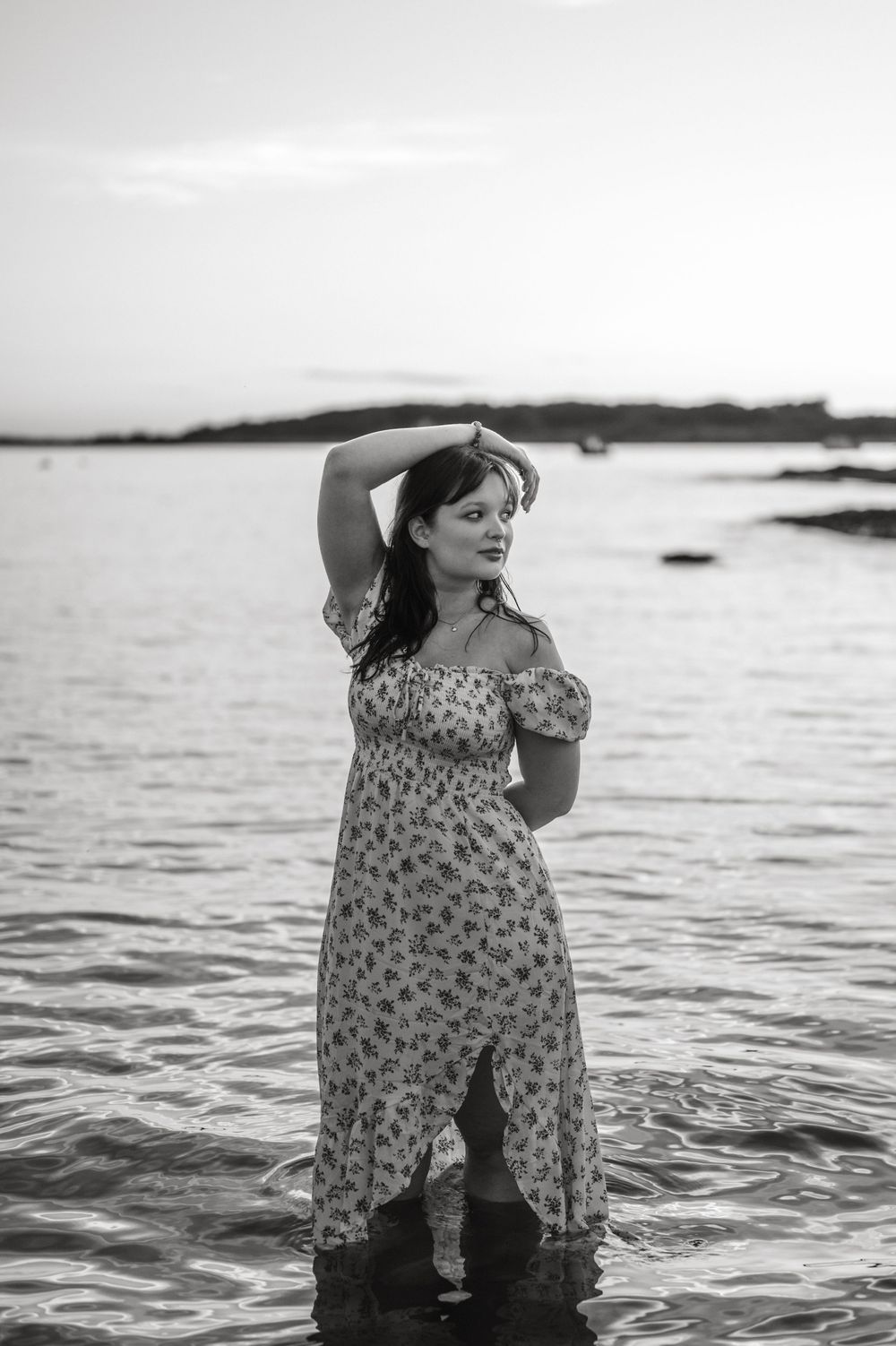 A dreamy black and white portrait of someone in a floral dress standing in shallow ocean waters.