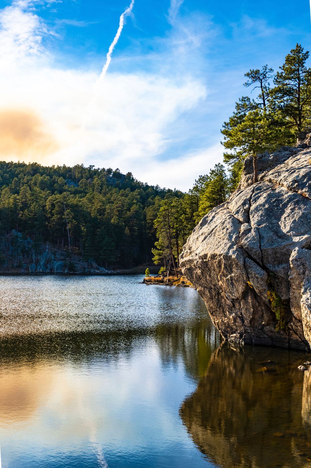 Dramatic granite cliff face jutting out over calm mountain lake surrounded by pine trees.