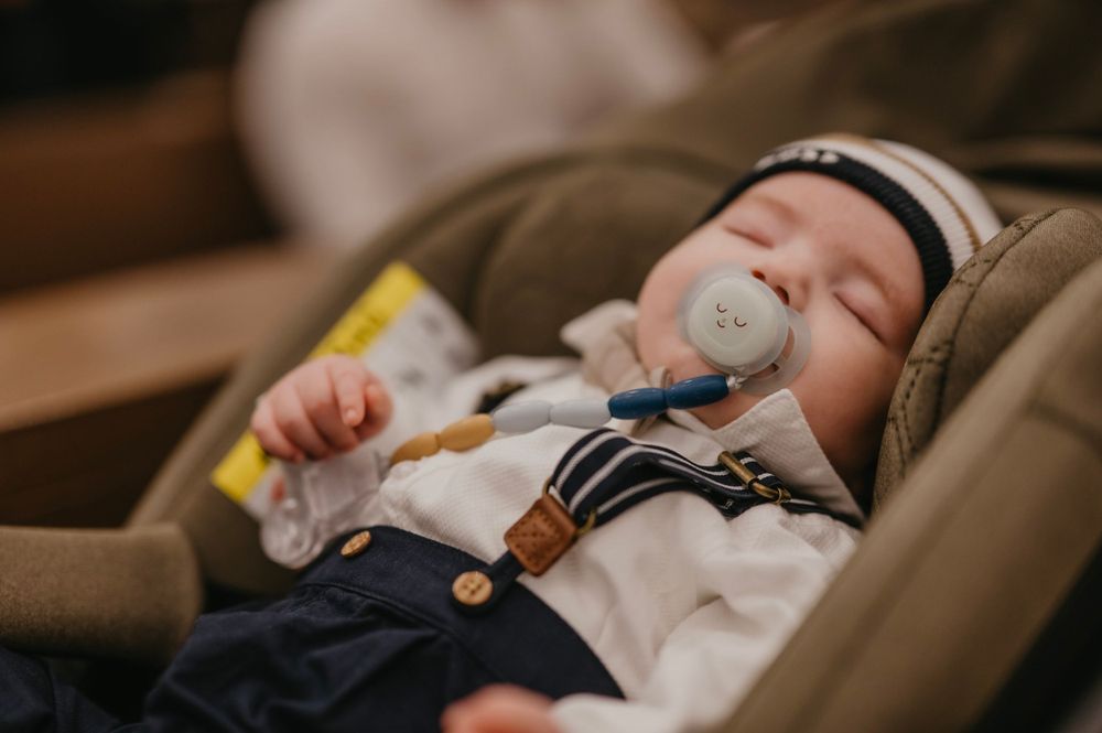 A sleeping baby in a car seat wearing a white outfit with navy shorts and a striped beanie hat.