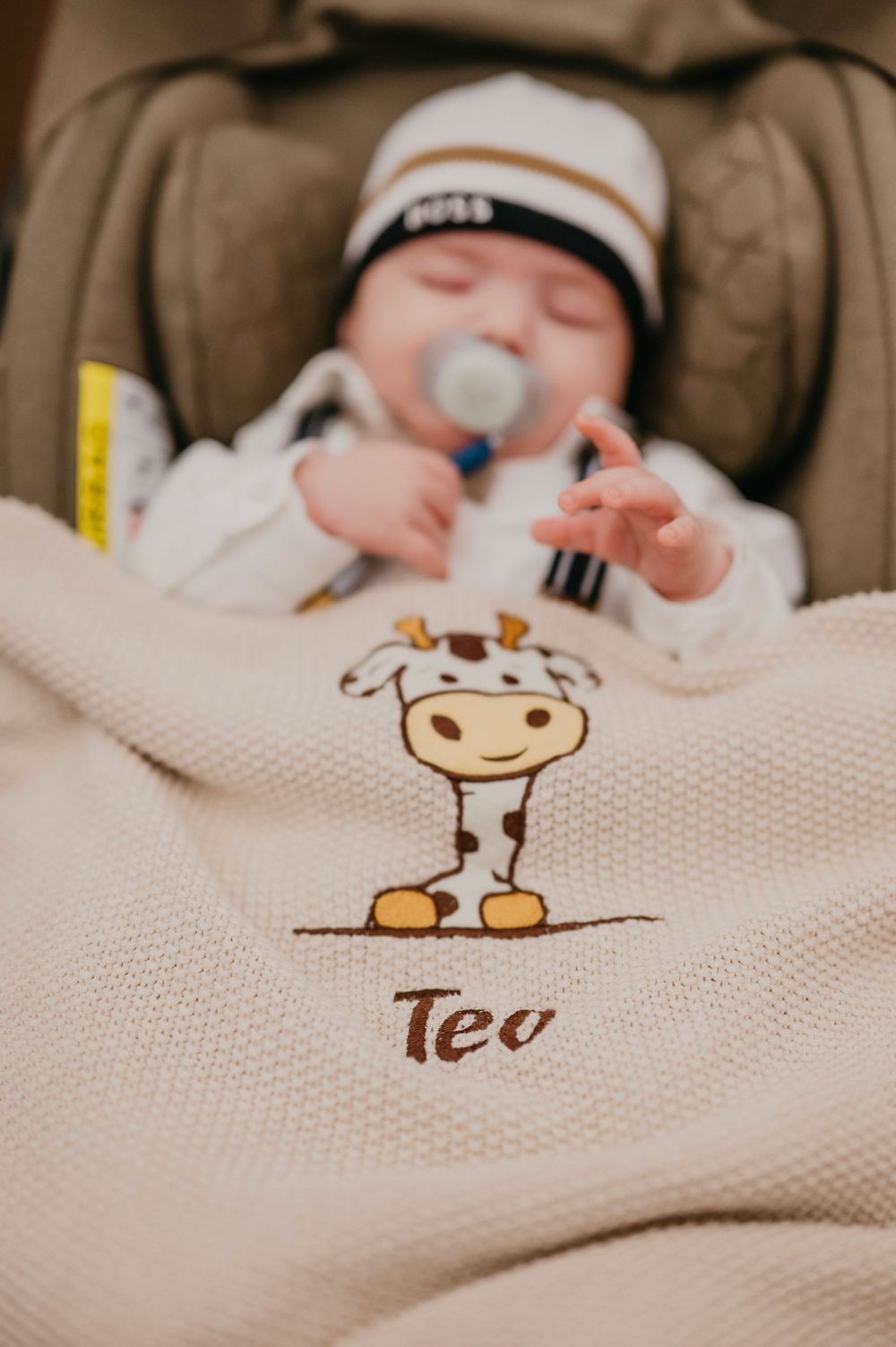 A baby sleeping in a car seat with a blanket featuring a cute giraffe design and 'Teo' text.