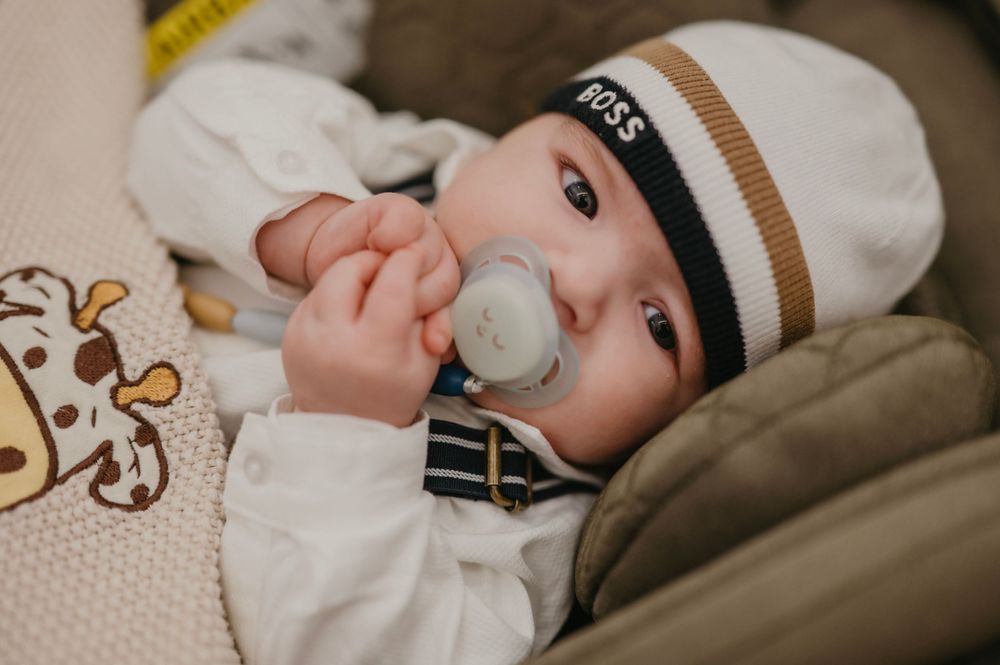 A baby wearing a striped beanie and white outfit lies contentedly in a car seat with a pacifier.