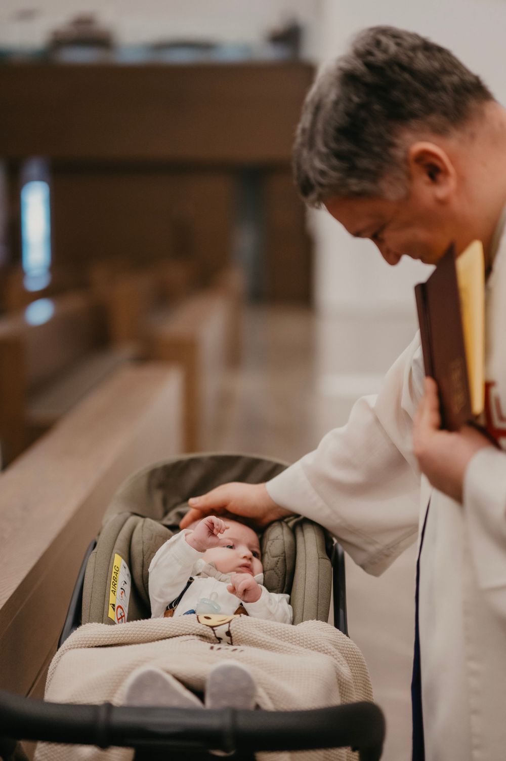 A priest in white robes leans over to bless a small baby in a car seat.